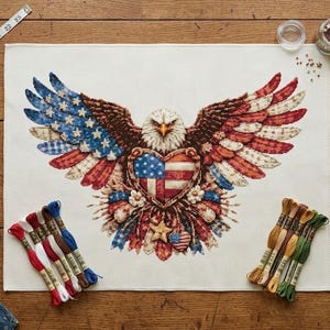 May include: A patriotic cross-stitch design featuring an American bald eagle with outstretched wings. The eagle's wings are colored with the American flag's stars and stripes. Embroidery floss, scissors, and a measuring tape are also visible.