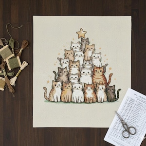 May include: A cross-stitch design depicting a Christmas tree formed by numerous cat illustrations. The cats are in shades of brown, white, and grey, with a gold star at the top. The design is on a beige fabric.