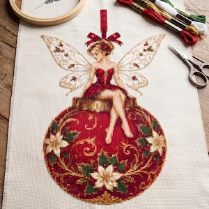 May include: A cross-stitch project on cream fabric featuring a fairy with translucent wings sitting on a large red Christmas bauble decorated with poinsettias. Includes a wooden embroidery hoop, a needle threader, four skeins of embroidery thread in red, gold, white, and green, and a pair of ornate metal scissors.