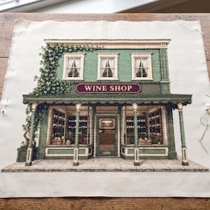 Peut inclure: Une broderie au point de croix sur tissu blanc représentant une devanture de magasin verte avec le texte WINE SHOP. Le bâtiment comporte trois fenêtres à l'étage, un porche couvert avec des colonnes et de grandes vitrines remplies de bouteilles.