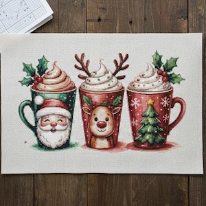 Peut inclure: Motif de point de croix représentant trois tasses festives avec de la crème fouettée et des motifs de Noël : le Père Noël, un renne et un sapin. Les tasses sont rouges et vertes, avec des accents de houx. L'image est sur un tissu crème.
