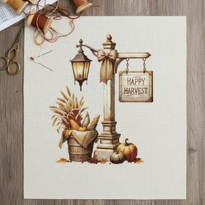 Peut inclure: Motif de point de croix représentant un lampadaire avec une enseigne "Happy Harvest". Un seau en bois déborde de maïs et de blé. Des citrouilles et des feuilles d'automne sont à la base. Des bobines de fil et des ciseaux sont en arrière-plan.