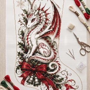 Op de afbeelding: Een kruissteekproject op witte stof met een witte draak met rode accenten, omringd door hulstbladeren, bessen en een grote rode strik met zilveren belletjes. Inclusief borduurgaren in rood, groen en crème, naalden en een schaar.