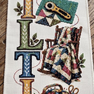 May include: A vertical cross-stitch sampler on cream fabric featuring the word QUILTING spelled out in large, colorful, patterned letters. Each letter is accompanied by embroidered motifs including fabric stacks, a rotary cutter, a rocking chair with a quilt, and a sewing basket filled with supplies.