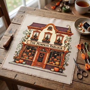 Puede incluir: Un proyecto de bordado en punto de cruz sobre tela beige que representa una tienda titulada The Pumpkin Parlour. El edificio cuenta con un techo oscuro, tres ventanas superiores con cortinas y grandes escaparates llenos de calabazas naranjas.