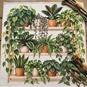 May include: A cross-stitch embroidery pattern on white fabric featuring a three-tiered wooden shelf filled with various potted houseplants, including trailing vines, ferns, and broad-leafed plants, accompanied by embroidery floss and scissors.