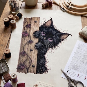 Puede incluir: Un proyecto de punto de cruz sobre tela blanca con un gatito negro esponjoso de grandes ojos morados asomándose tras un poste de madera con enredaderas. Una etiqueta indica PATTERN SIZE: 158x208 STITCHES, junto a hilos y unas tijeras.
