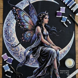 Peut inclure: Un motif de point de croix sur tissu noir représentant une fée aux ailes irisées assise sur un croissant de lune. Le design inclut des étoiles et des motifs ornés. Bobines de fil, ciseaux, aiguille et règle indiquant 61,0 cm x 71,1 cm.