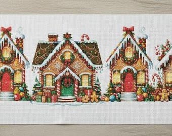 May include: A cross-stitch project featuring five gingerbread houses with snow-covered roofs, wreaths, and festive decorations. The houses are in a row on a white background. The scene includes a cup of coffee, embroidery supplies, and scissors.
