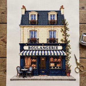 Könnte beinhalten: Eine Kreuzstichstickerei eines französischen Bäckereigebäudes mit dem Wort BOULANGERIE auf der Ladenfront. Das Design zeigt eine blau-weiß gestreifte Markise, zwei kleine Stühle mit einem Tisch und eine Kletterpflanze an der rechten Fassadenseite.