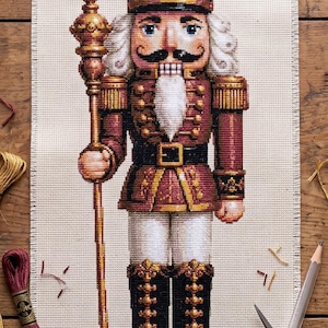 Peut inclure: Une broderie au point de croix représentant un soldat casse-noisette portant un chapeau noir, une veste rouge avec des bordures dorées et des bottes noires. La figurine tient un grand bâton doré et se tient sur une base circulaire.