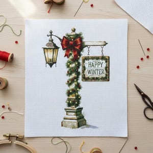 May include: Cross-stitch design depicting a winter scene with a lamppost decorated with a red bow, garland, and a sign reading "Happy Winter". The design is on white fabric, with sewing supplies around it.