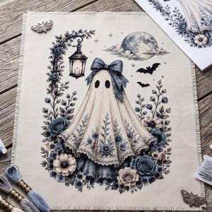 May include: A cross-stitch project on beige fabric featuring a central ghost figure wearing a blue bow, surrounded by blue and cream floral arrangements, a hanging lantern, a full moon, and two bats, accompanied by embroidery floss and scissors.