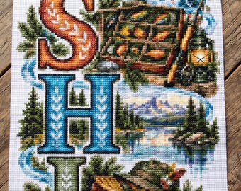 May include: A vertical cross-stitch embroidery on white Aida fabric featuring the word FISHING in large, colorful block letters. Each letter incorporates detailed outdoor scenes including a cabin, a jumping trout, a lantern, and a waterfall.