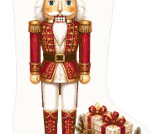 Op de afbeelding: Een witte kerstsok met een opdruk van een notenkraker-soldaat in een rood en goud uniform met een bijpassende kroon. Aan de voet van de notenkraker liggen verschillende ingepakte cadeautjes, gouden en rode kerstballen en dennentakken.