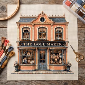 May include: A cross-stitch pattern on fabric depicting a two-story shop titled THE DOLL MAKER. The building features peach-colored walls, a dark blue roof, and large display windows filled with miniature dolls. Embroidery floss and scissors are included.