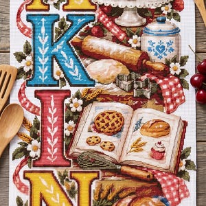 Może przedstawiać: Prostokątny biały ręcznik z tkaniny z haftem krzyżykowym przedstawiającym słowo BAKING dużymi, kolorowymi literami. Obraz zawiera niebieski mikser stojący, babeczki, wałek do ciasta, książkę kucharską, chleb i ciasto, otoczone kwiatami.