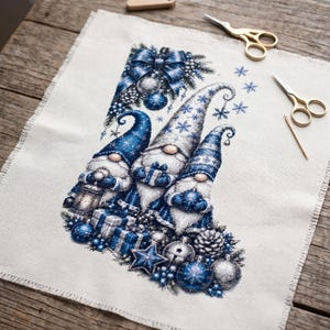 May include: A cross-stitch embroidery on off-white fabric featuring three gnomes in blue and white hats, surrounded by blue Christmas ornaments, pinecones, and gift boxes. The design includes a large blue bow at the top and several snowflake motifs.