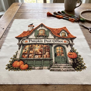 Puede incluir: Un bordado de punto de cruz sobre tela blanca que representa un edificio con la etiqueta Pumpkin Post Office. El diseño presenta un tejado naranja, una fachada verde, calabazas y un pájaro negro en la chimenea. Hilos, bastidor y tijeras están cerca.