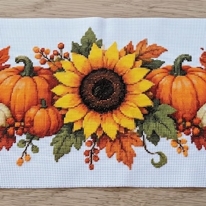 Puede incluir: Un camino de mesa rectangular blanco con un patrón central de punto de cruz que muestra tres grandes girasoles amarillos, dos calabazas naranjas, dos calabazas blancas pequeñas y varias hojas de arce otoñales y bayas rojas.
