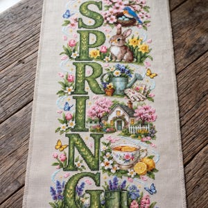 May include: A rectangular linen fabric piece featuring a vertical cross-stitch design with the word SPRING in large green letters. The embroidery includes a rabbit, a bird in a nest, a watering can, a house, a teacup with lemons, and green boots.