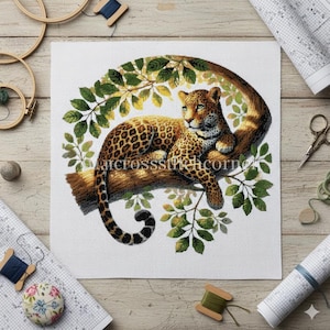 May include: Cross-stitch of a leopard resting on a tree branch, surrounded by green leaves. The leopard has a spotted coat and green eyes. The artwork is on white fabric, with sewing supplies around the edges.