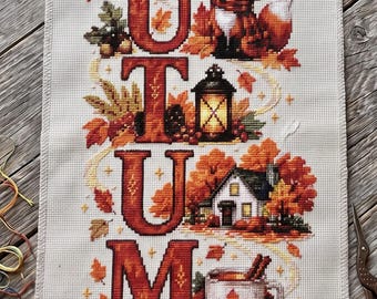 May include: A vertical cross-stitch embroidery on light beige fabric featuring the word AUTUMN in large red letters. The design includes a pumpkin, a fox, a lantern, a house, a mug with cinnamon sticks, and boots, all surrounded by colorful fall leaves.