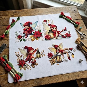 May include: Cross-stitch project featuring four panels with fairy illustrations in red outfits, holding lanterns, and surrounded by poinsettias. Embroidery floss, needles, and scissors are arranged around the fabric on a wooden surface.