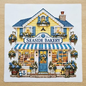May include: A cross-stitch embroidery pattern on white fabric depicting a yellow two-storey building with a blue roof and striped awning. The sign reads SEASIDE BAKERY. The scene includes blue shutters, flower boxes, hanging baskets, and a blue door.