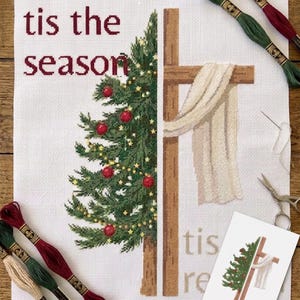 May include: A cross-stitch design with a Christmas tree and a cross. The text "tis the season" is stitched in burgundy. The tree is decorated with red baubles and yellow lights. Embroidery threads in green, burgundy, and beige are visible.
