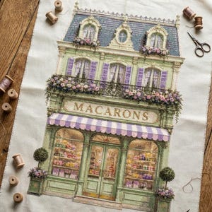 Può includere: Un ricamo a punto croce su tessuto bianco che raffigura una vetrina a più piani con la parola MACARONS stampata sopra l'ingresso. L'edificio presenta una facciata verde, persiane viola, una tenda a righe e fioriere alle finestre.