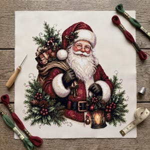 Puede incluir: Un patrón de punto de cruz sobre tela blanca que muestra a Santa Claus con un abrigo rojo con ribete de piel blanca, cargando un saco de regalos y una linterna. Rodeado de ramas de pino y bayas, con hilo de bordar, una aguja y una cinta métrica.