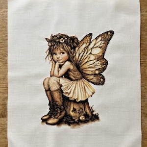 May include: Cross-stitch of a fairy with brown hair, a floral crown, and large, detailed wings. The fairy is seated on a tree stump, wearing a dress and boots. The artwork is on a white fabric background.