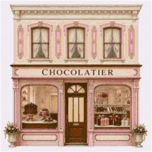 May include: A cross-stitch pattern of a two-storey chocolatier shop with a pale pink facade, brown central door, and large display windows. The storefront features the word "CHOCOLATIER" in dark lettering above the entrance and windows.