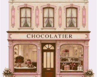 May include: A cross-stitch pattern of a two-story chocolatier shop with a pale pink facade, brown central door, and large display windows. The storefront features the word "CHOCOLATIER" in dark lettering above the entrance and windows.
