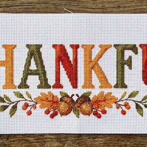 Könnte beinhalten: Ein weißes Stoffstück mit einem Kreuzstich-Design des Wortes THANKFUL in abwechselnd roten, orangefarbenen und grünen Buchstaben. Der Text wird von herbstlichen Motiven wie gelben Sonnenblumen, braunen Eicheln und roten Beeren umrahmt.