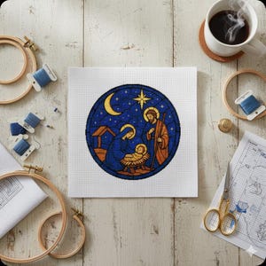 May include: A cross-stitch design depicting a nativity scene in a stained-glass style circle, with a crescent moon and star. The figures of Mary, Joseph, and baby Jesus are visible. Embroidery hoops, thread, and a coffee cup are arranged around the project.