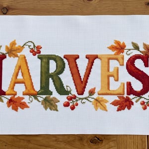 Puede incluir: Una pieza de tela rectangular blanca con un diseño de punto de cruz que muestra la palabra HARVEST en letras de bloque multicolores. El texto está flanqueado por calabazas naranjas, manzanas rojas, trigo dorado y hojas de otoño.