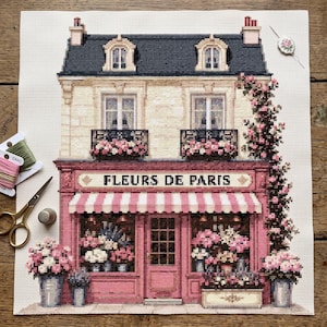 May include: A cross-stitch embroidery of a Parisian flower shop titled Fleurs de Paris, featuring a pink storefront with a striped awning, a dark roof, and climbing roses. The piece is displayed with gold scissors, a thimble, and thread spools.