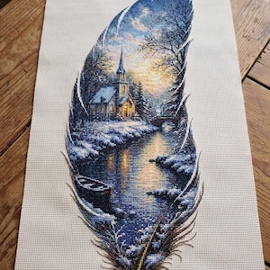May include: A rectangular piece of off-white Aida fabric featuring a detailed cross-stitch design shaped like a large feather. The interior of the feather depicts a winter landscape with a church, a snow-covered river, and a small boat at dusk.