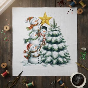 Op de afbeelding: Een kerst-thema kruissteekontwerp met een sneeuwmanfamilie, een gouden ster en een met sneeuw bedekte kerstboom. De sneeuwmannen dragen feestelijke hoeden en sjaals. Naaibenodigdheden, zoals garenklosjes en scharen, omringen de afbeelding.