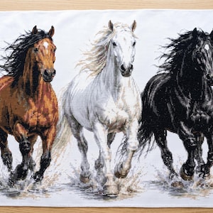 Puede incluir: Una pieza de tela rectangular blanca con un bordado de punto de cruz detallado de tres caballos corriendo por el agua. Los caballos están representados en marrón, blanco y negro, con crines y colas fluidas, capturados al galope.