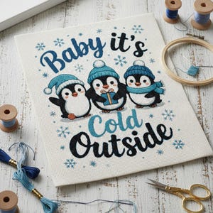 May include: Cross-stitch artwork featuring three penguins wearing blue hats and scarves, with the text "Baby it's Cold Outside." The penguins are holding a gift. The artwork is surrounded by snowflakes. Sewing supplies, including thread, scissors, and a hoop, are visible.
