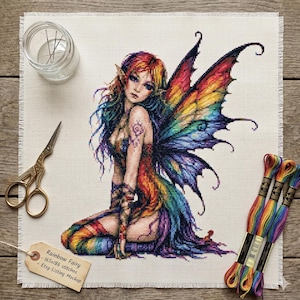 May include: A cross-stitch pattern on white fabric featuring a fairy with rainbow-coloured hair, wings, and clothing. The kit includes gold-toned embroidery scissors, a clear needle holder, and three skeins of rainbow-coloured embroidery floss.