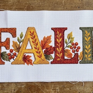 May include: A white Aida fabric piece featuring a cross-stitch design of the word FALL in large, decorative letters. The letters are surrounded by embroidered sunflowers, red berries, and autumn leaves in shades of orange, yellow, and green.