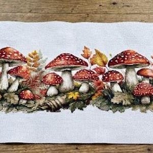 May include: A cross-stitch project featuring a vibrant mushroom design with red caps and white spots, surrounded by autumn leaves. The scene includes embroidery floss, scissors, and an embroidery hoop, all arranged on a rustic wooden surface.