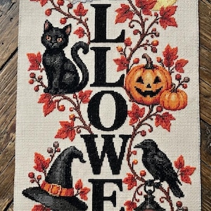 Puede incluir: Un muestrario de punto de cruz vertical sobre tela beige con la palabra HALLOWEEN en letras negras grandes. El diseño incluye un fantasma blanco, una luna amarilla con un murciélago negro, un gato negro, calabazas naranjas, un sombrero de bruja, un cuervo y una linterna, rodeados de hojas de otoño rojas y naranjas. Unas tijeras de metal y tres madejas de hilo están cerca.