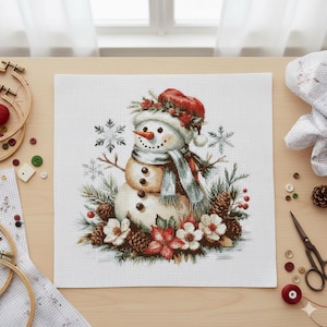 May include: Cross-stitch design featuring a snowman wearing a red hat and scarf, surrounded by snowflakes, pinecones, and flowers. The snowman has a carrot nose and button eyes. The design is on a white fabric background.