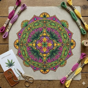 May include: A completed cross-stitch mandala with a cannabis leaf design in shades of purple, green, and yellow. The project is surrounded by spools of embroidery floss in various colors, scissors, and a pattern snippet on a wooden surface.