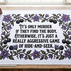May include: White fabric with embroidered text: "It's only murder if they find the body. Otherwise, it's just a really aggressive game of hide-and-seek." The text is framed by purple flowers and black berries. The fabric is on a wooden table.